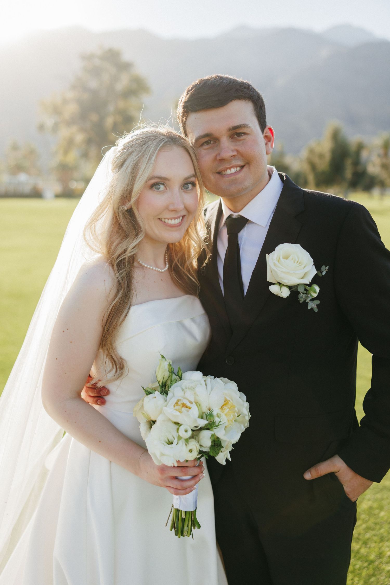 Smiling bride and groom pose together outdoors, bride holding white rose bouquet with mountains behind.