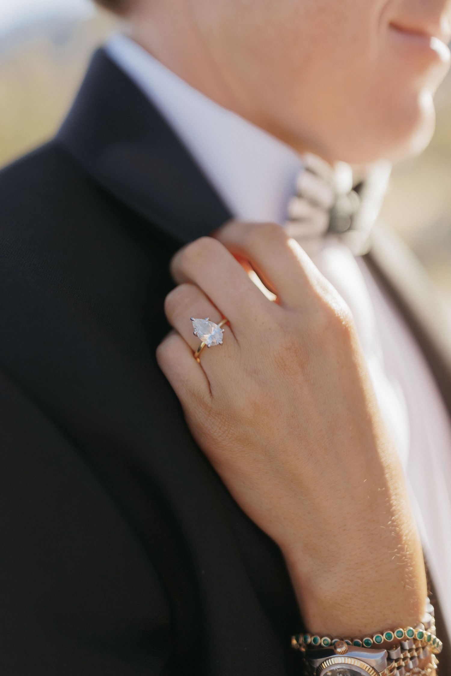 Close-up of brides pear-shaped diamond engagement ring as she rests her hand on grooms black tuxedo lapel.