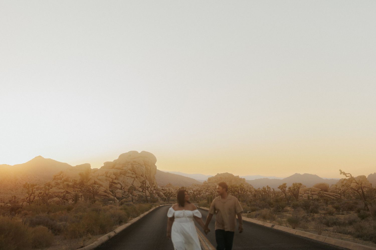 Couple in wedding attire walking down desert road at golden sunset with rocky mountains in Joshua Tree.