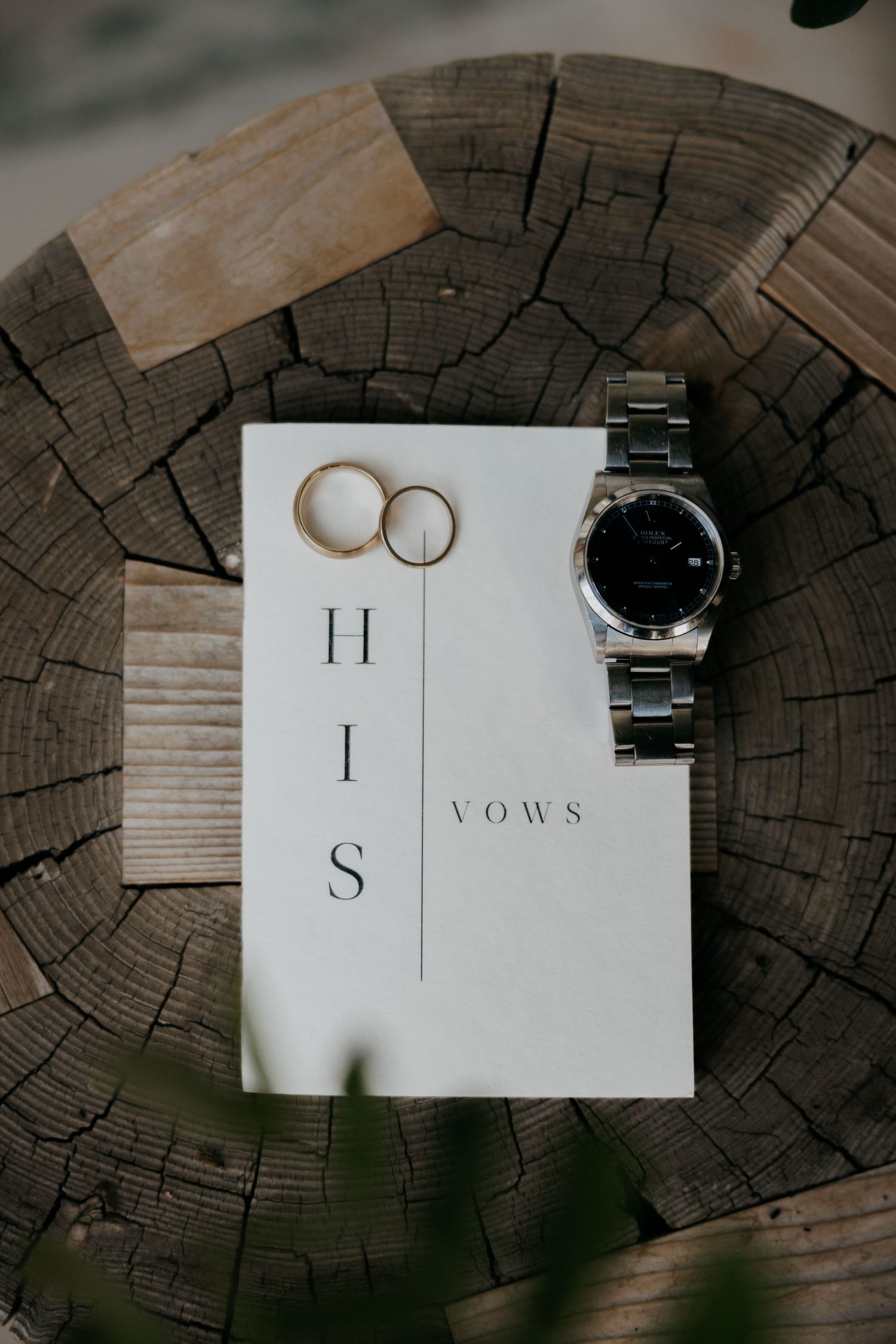 Wedding flat lay with his vows booklet, two gold rings, and a silver watch on a wooden stump.
