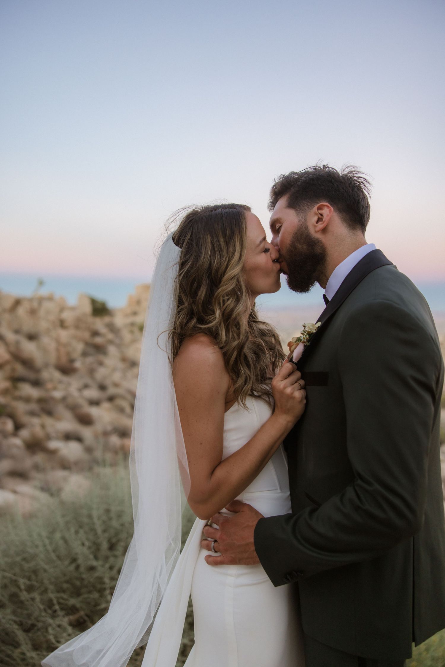 Romantic newlywed couple sharing intimate kiss moments at sunset in desert landscape with mountain scenery.