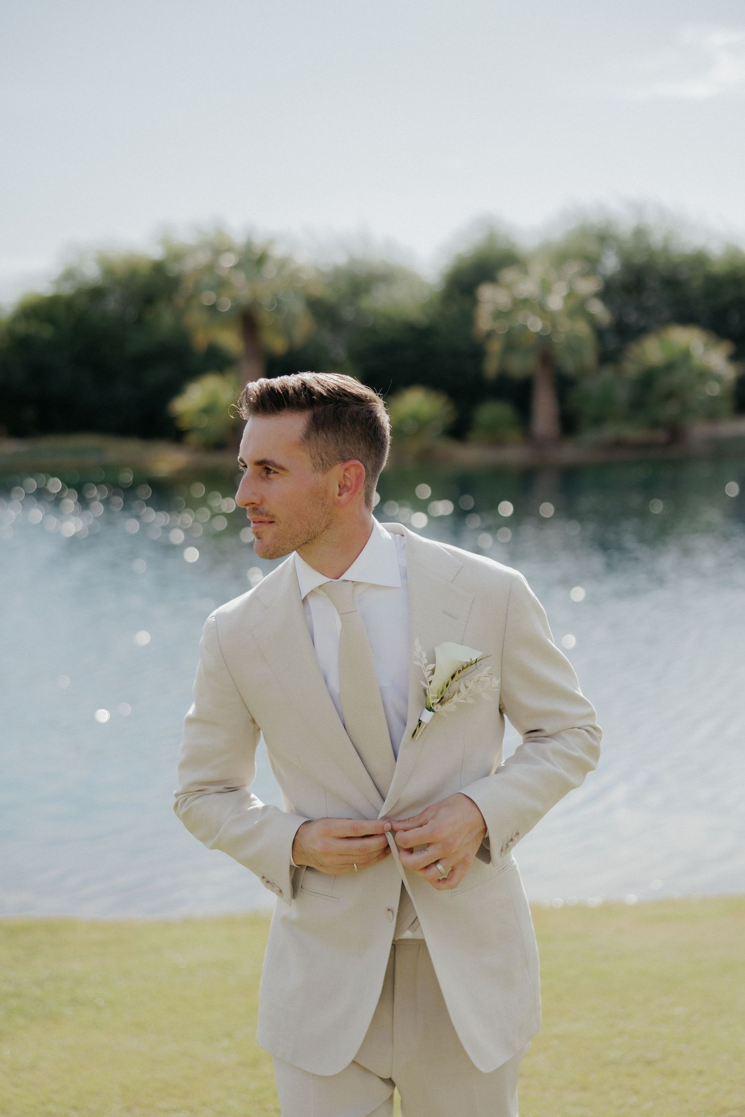 A person wearing a light beige suit poses gracefully on a grassy lawn beside a beautiful lake on a sunny day.