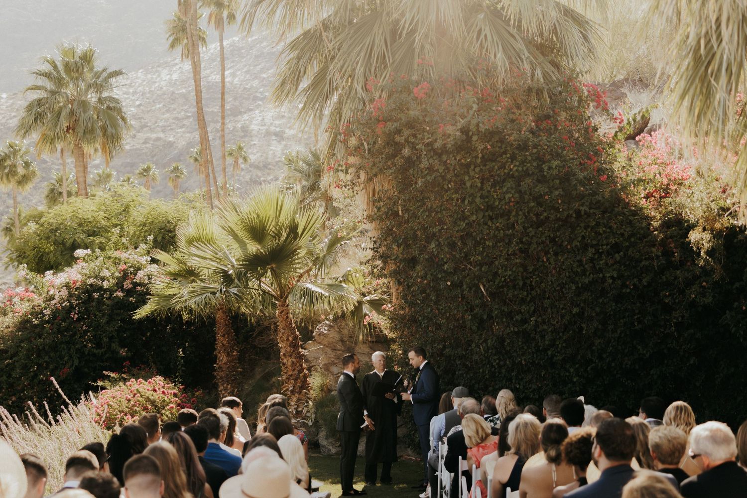 An outdoor garden wedding ceremony takes place under swaying palm trees during golden hour.