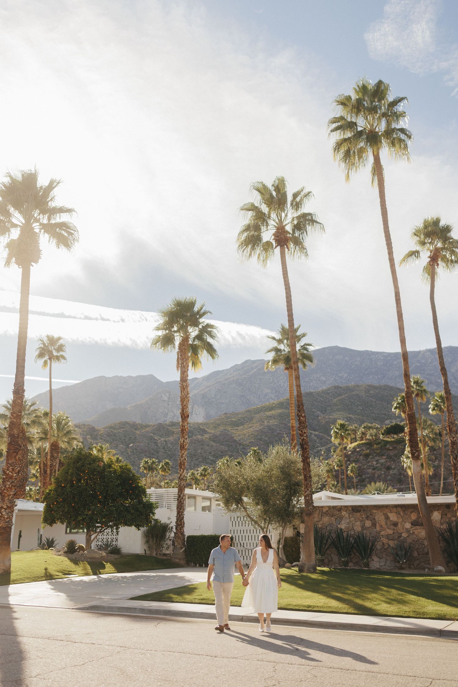 Couple holding hands walking among tall palm trees with mountain backdrop in sunny Palm Springs, CA.