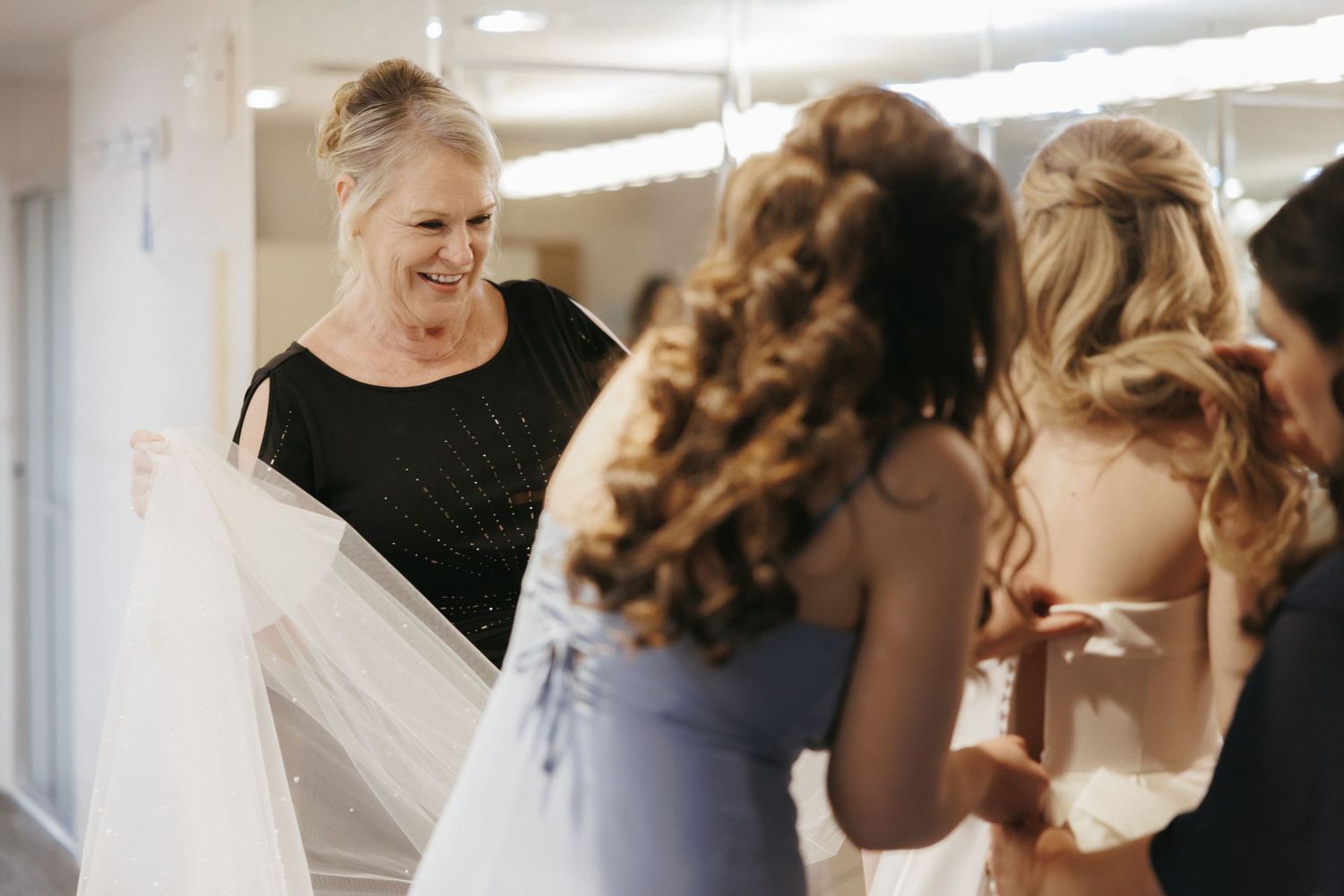 Smiling woman helping bride with wedding veil while bridesmaids assist with gown in a bright dressing room.