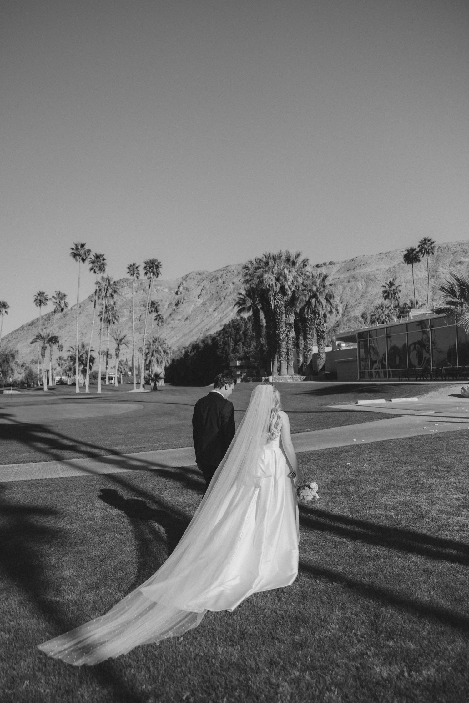 Bride and groom walk hand in hand with palm trees and mountains in black and white.