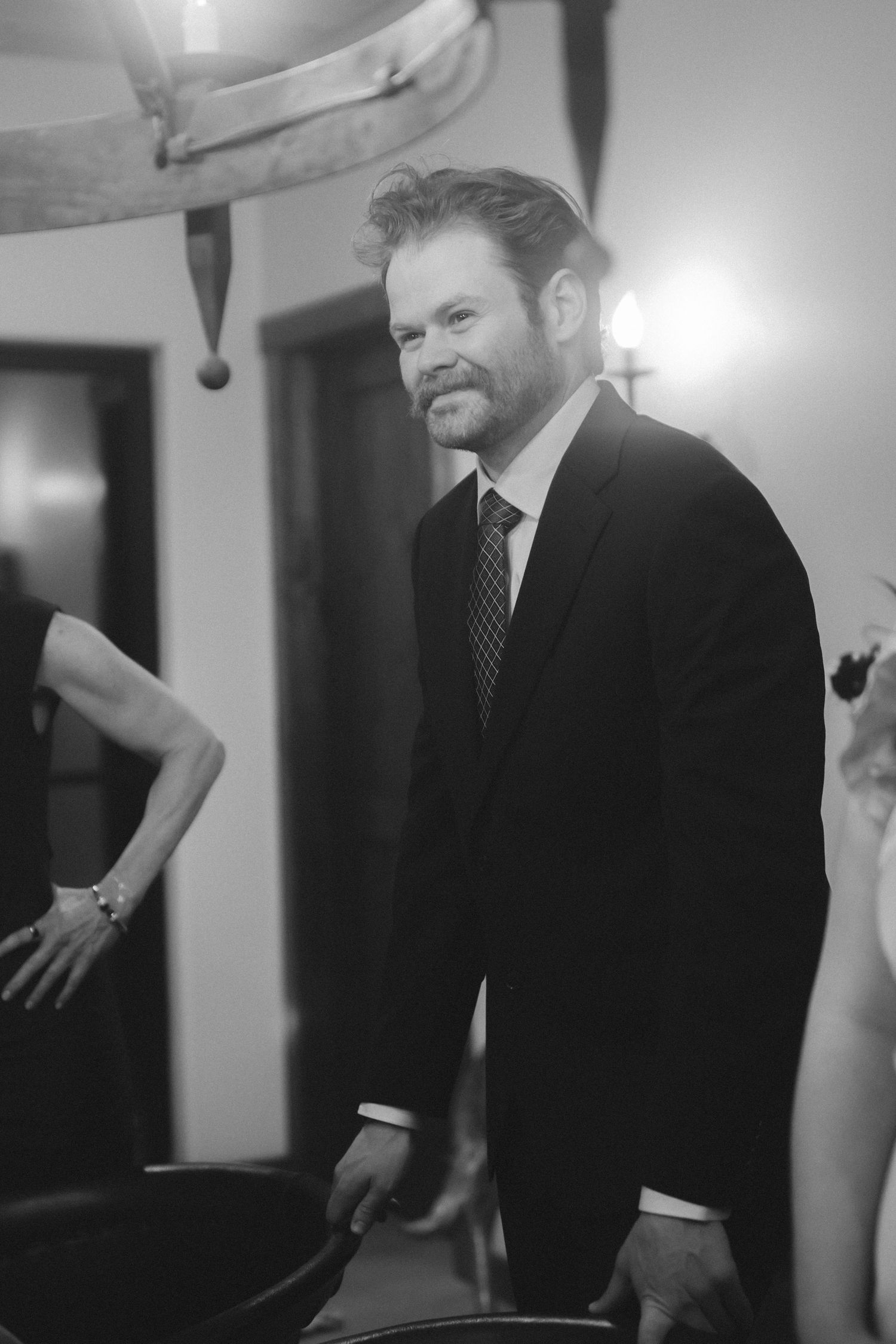 A man in a dark suit smiles warmly at a dimly lit indoor wedding reception.