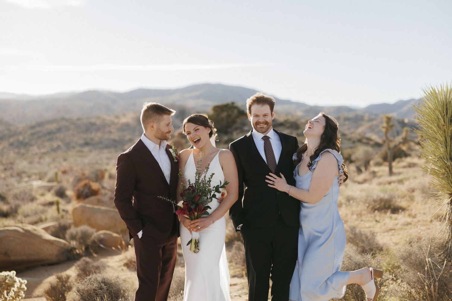 Wedding party of four laughs together in sunny Joshua Tree desert with mountain views behind them.