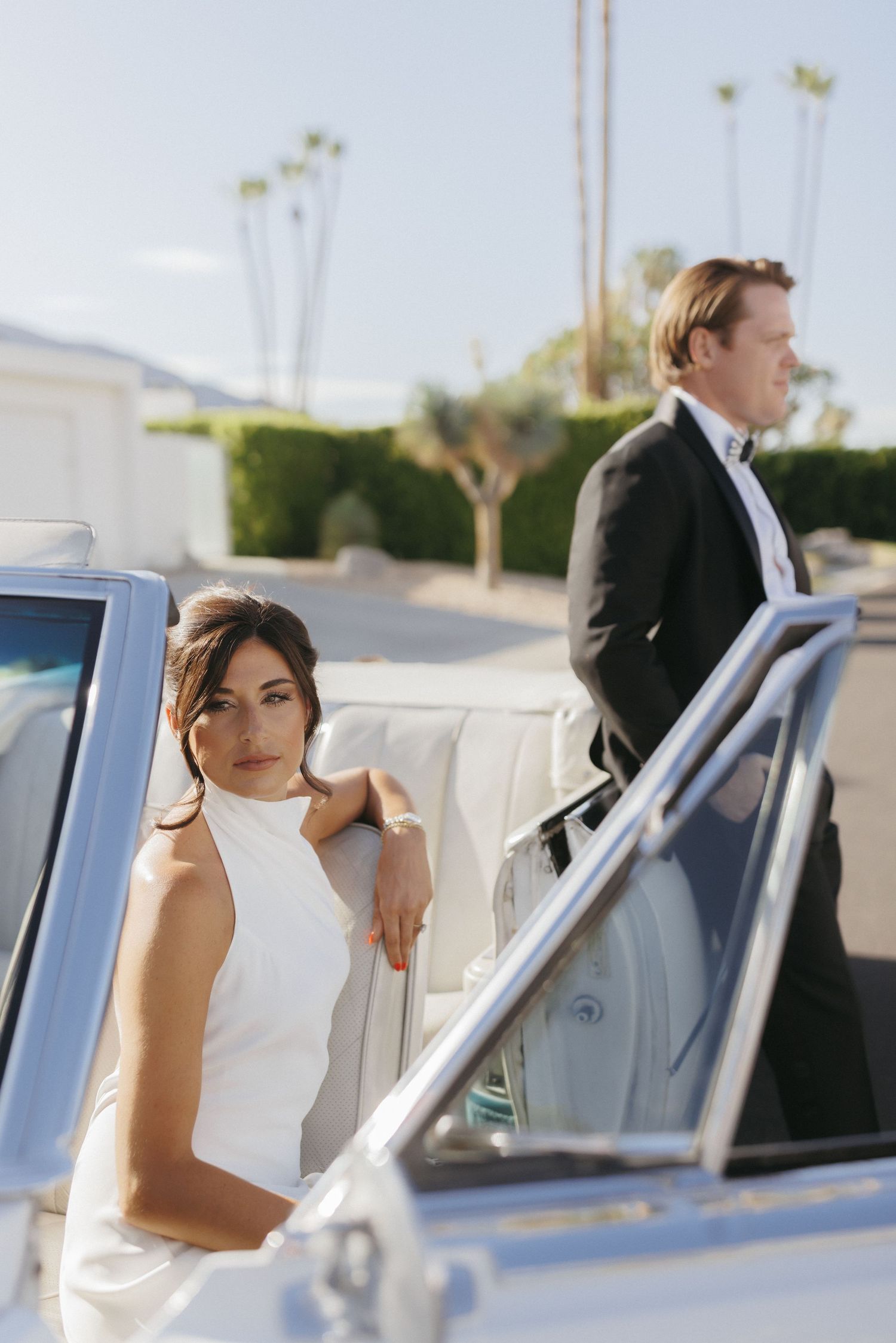 Bride in white halter gown leans on vintage white convertible with groom in tuxedo standing behind in Palm Springs.