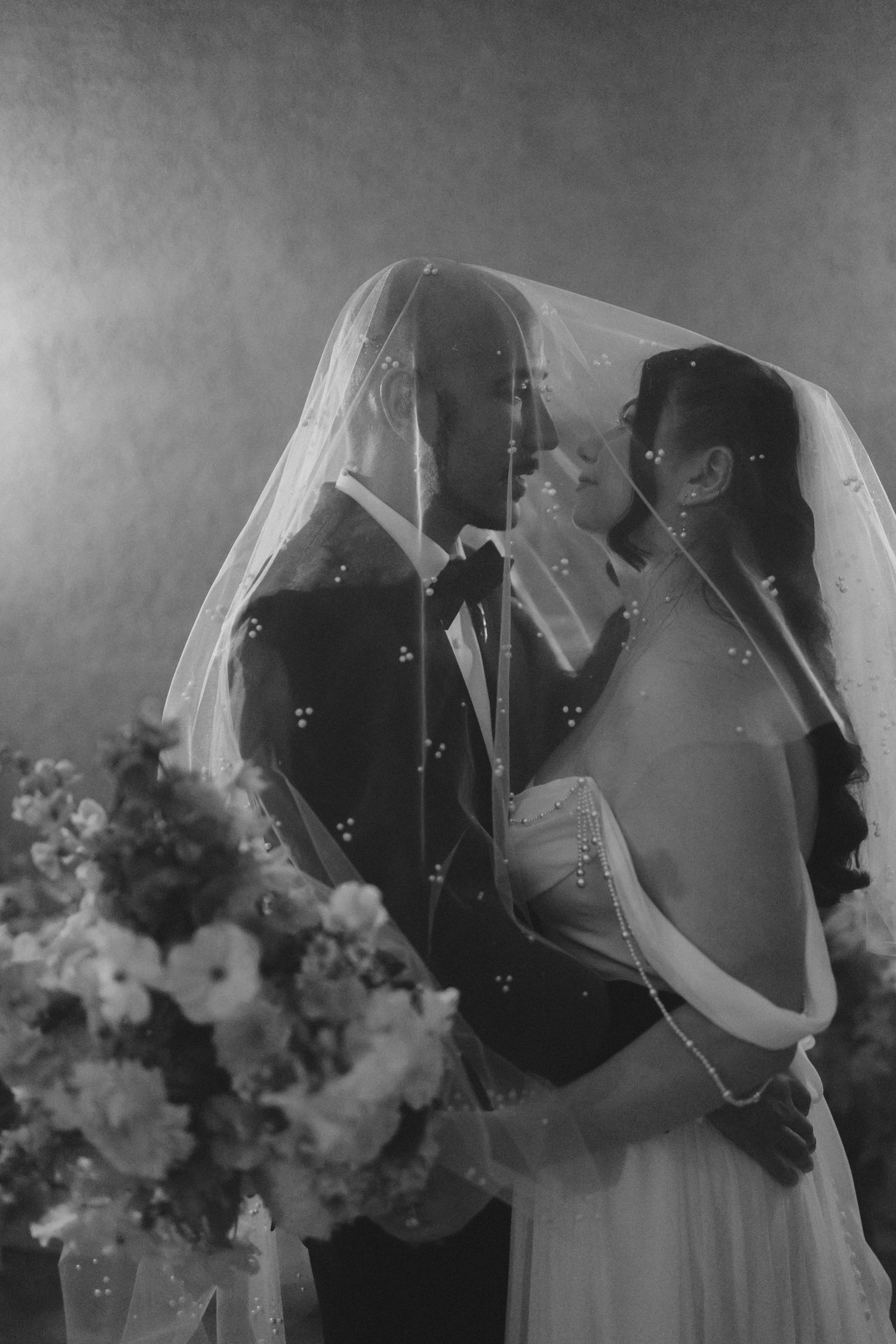 Black and white photo of bride and groom sharing intimate moment under wedding veil with floral arrangement nearby.