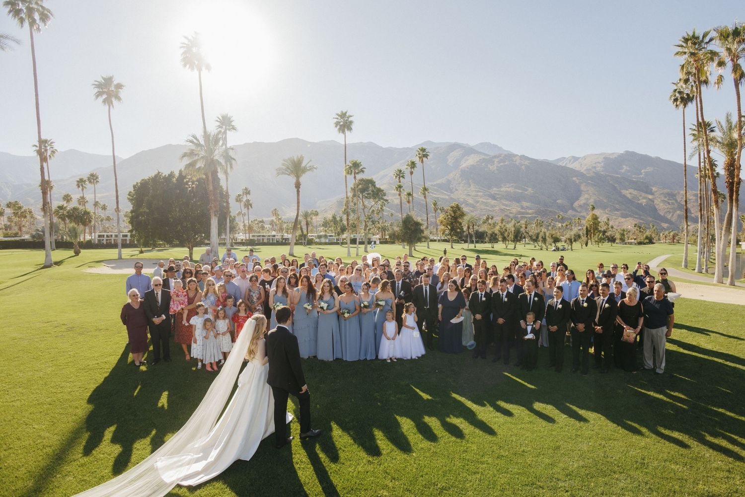 Aerial view of bride, groom, and large wedding party posing on a golf course with palm trees and mountains.