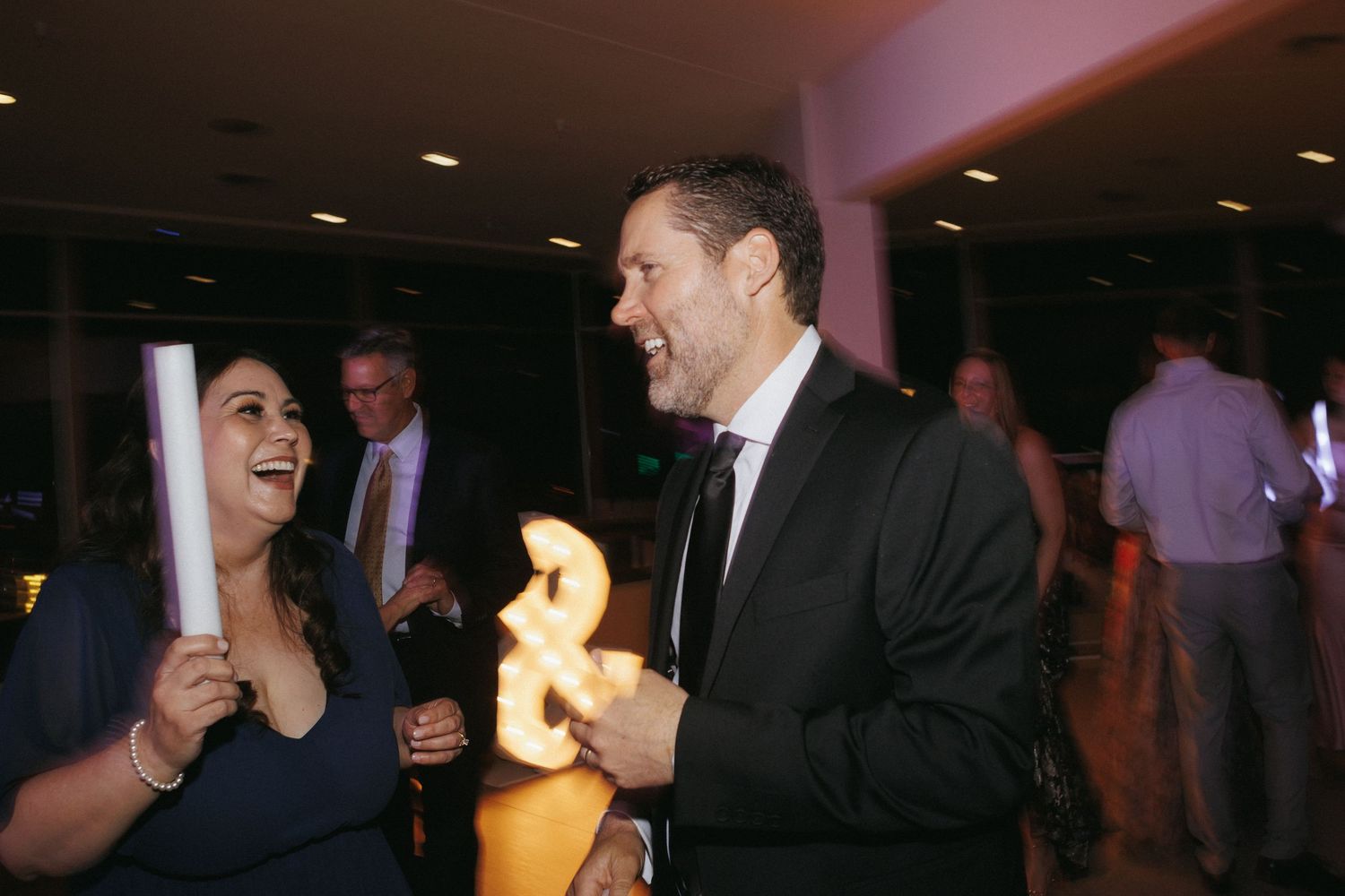 Guests laugh and celebrate on wedding reception dance floor holding glowing ampersand sign.