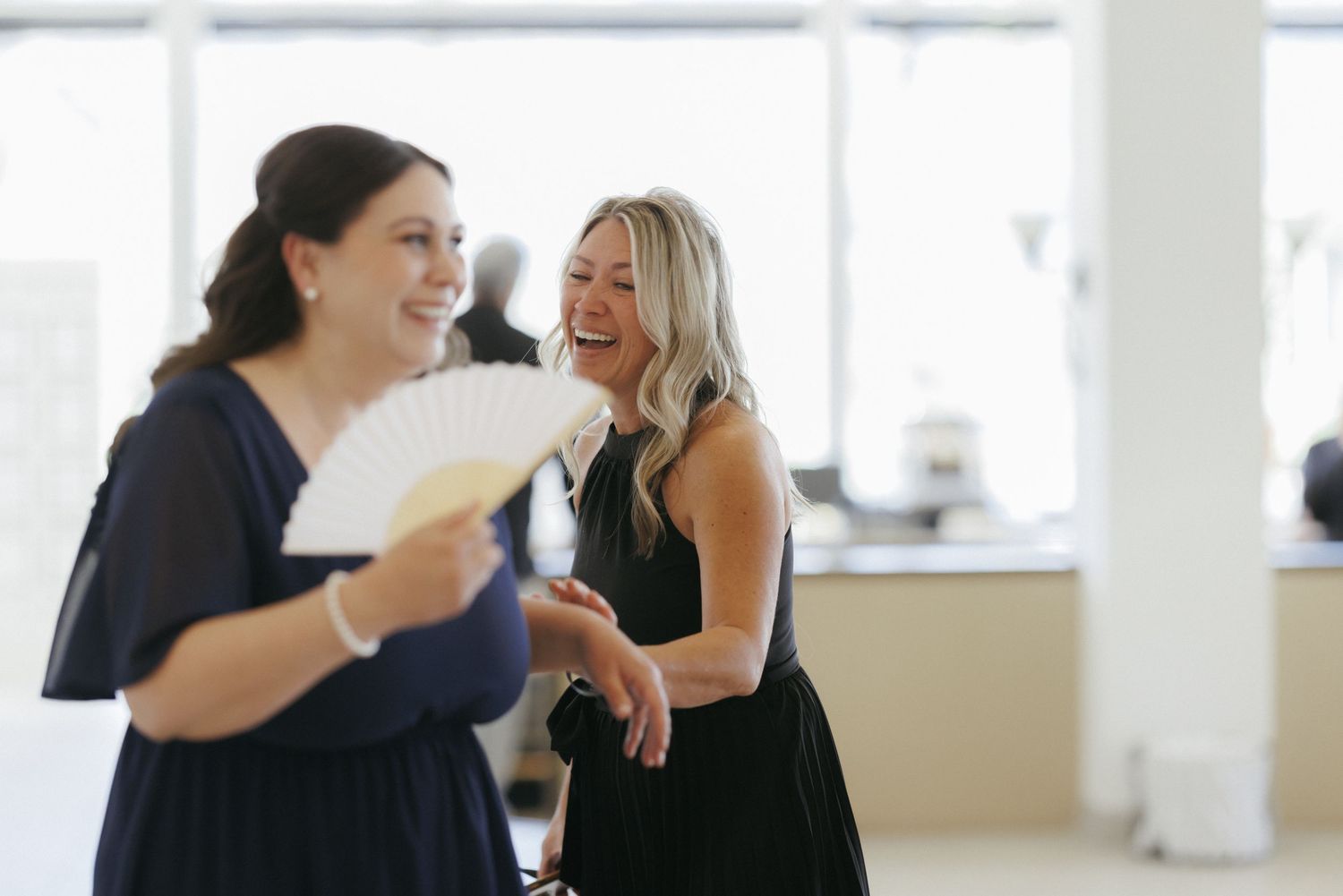 Two women in navy and black dresses laughing together, one holding a white hand fan at a wedding.