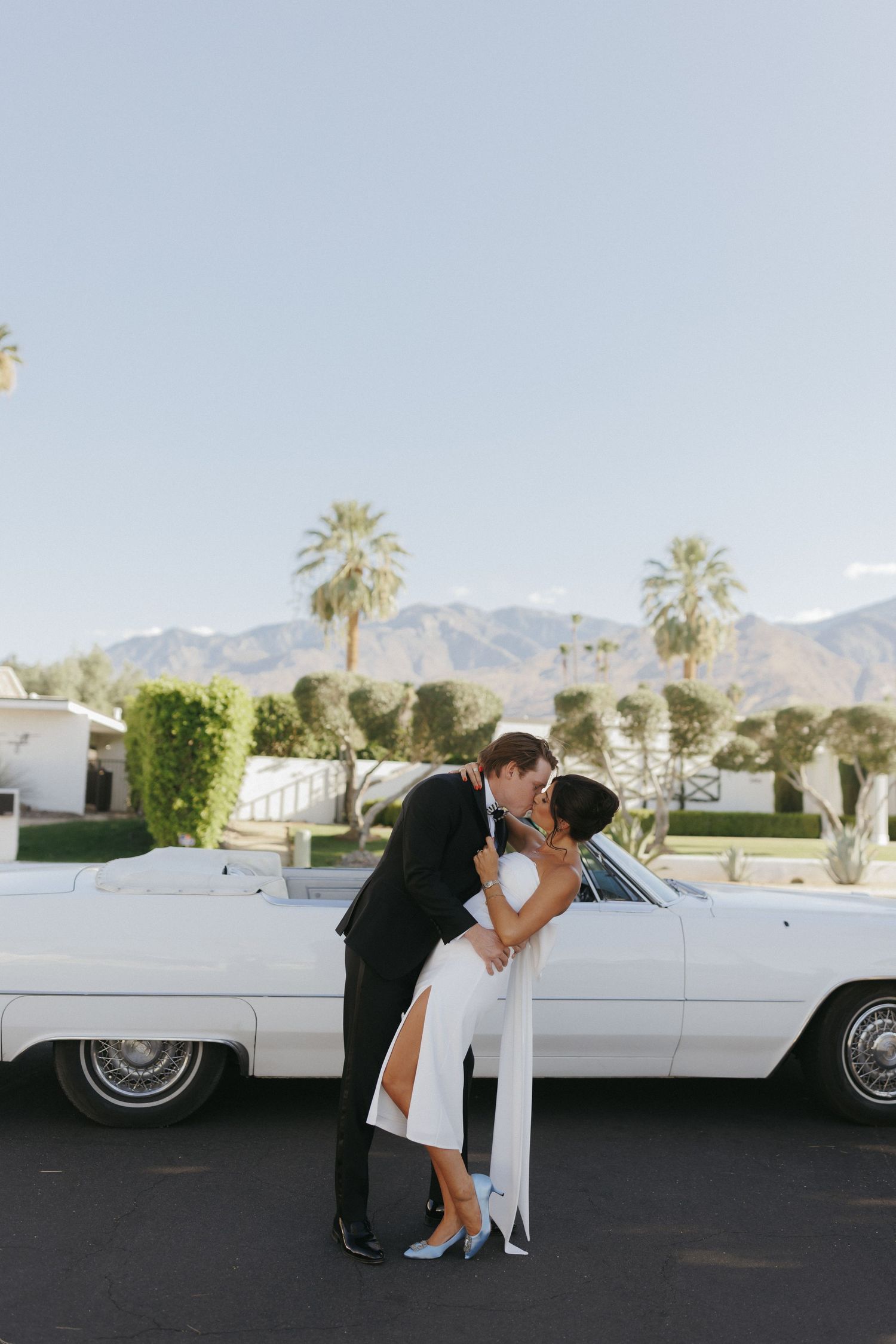 Bride and groom share a romantic kiss in front of a white vintage convertible with palm trees and mountains in Palm Springs.