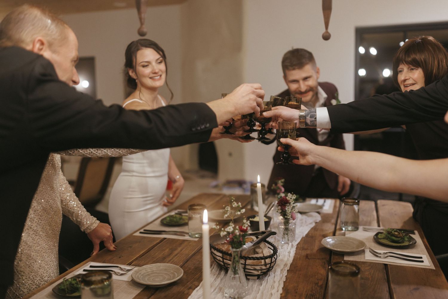 Wedding guests clink glasses in celebratory toast around candlelit rustic wooden reception dinner table.