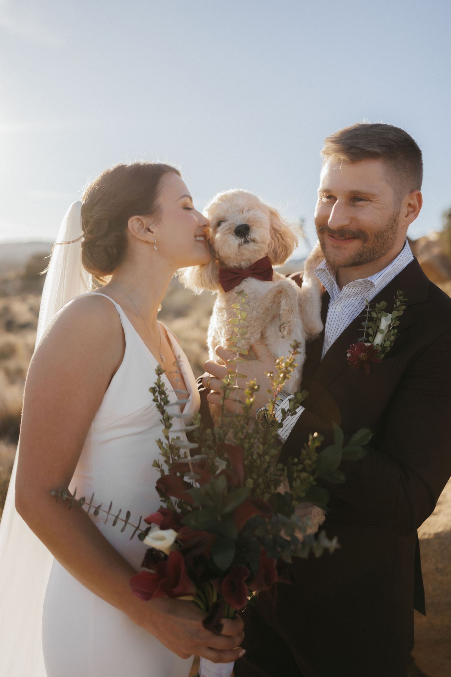 Smiling bride and groom hold adorable white dog wearing bow tie at desert wedding ceremony.