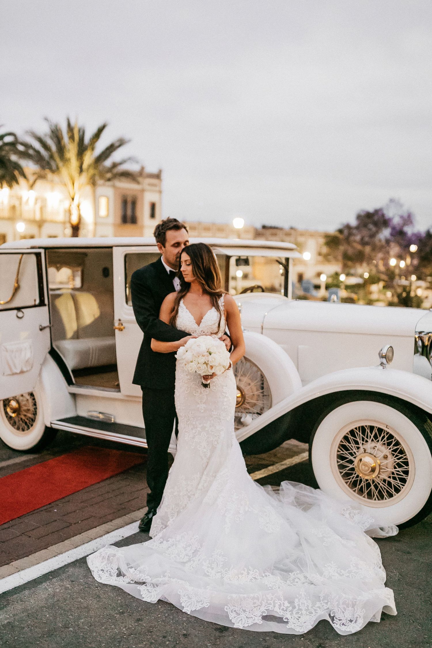 A couple shares a romantic moment next to a vintage white classic car during sunset at their wedding celebration.