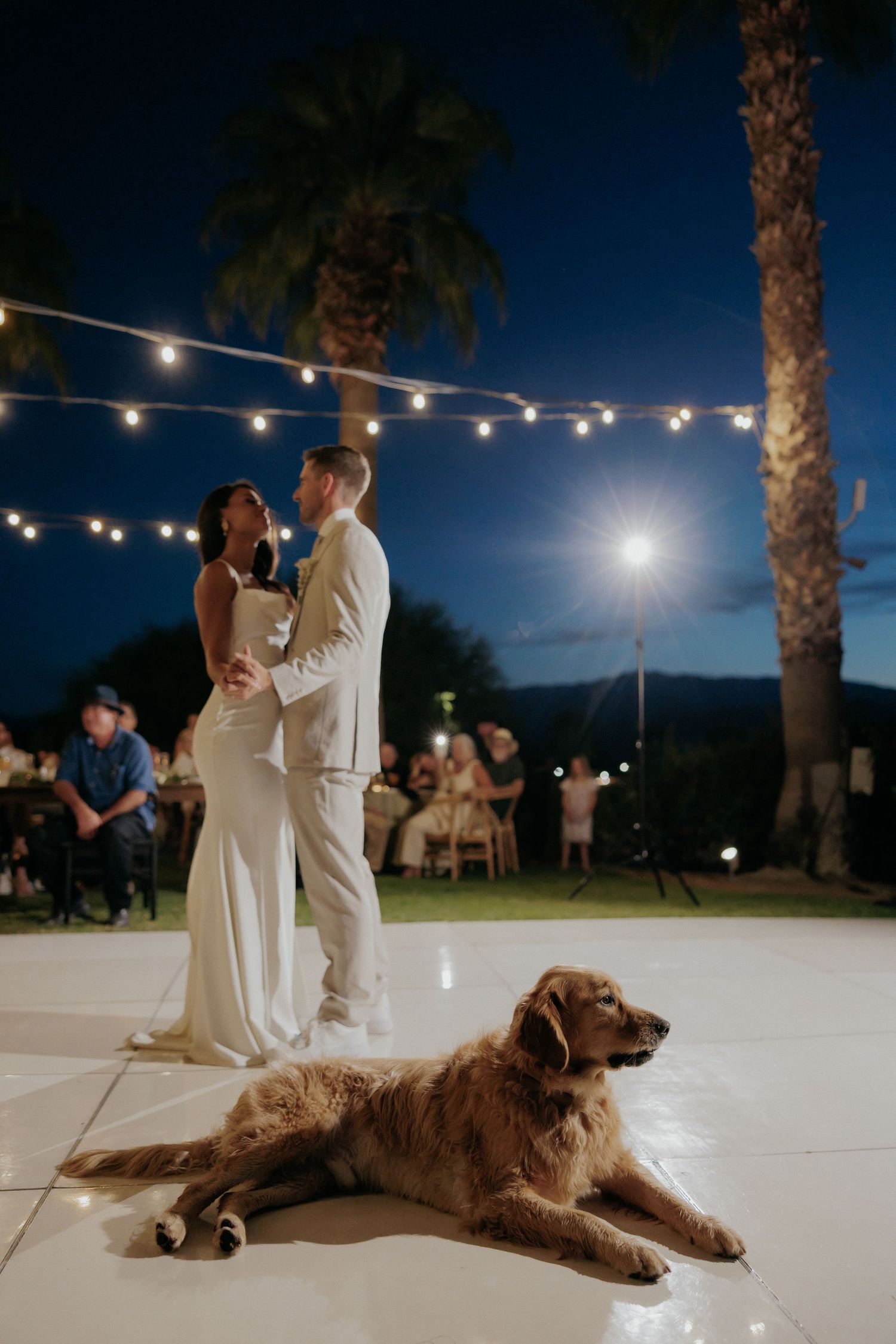A couple dances at an outdoor wedding reception with their dog resting on the dance floor under string lights at night.