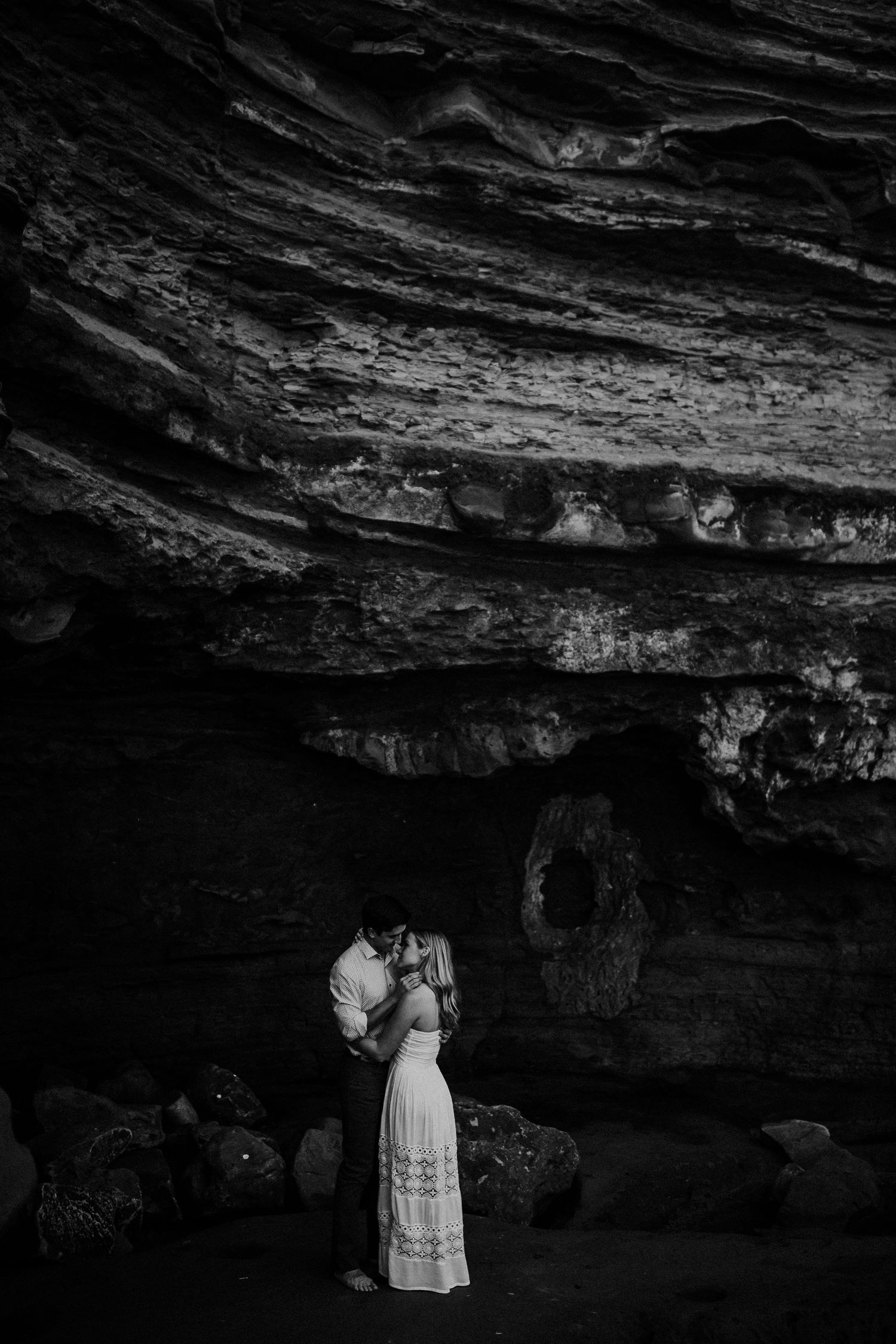 A romantic wedding couple stands together under a dramatic rock cave overhang with textured stone walls.