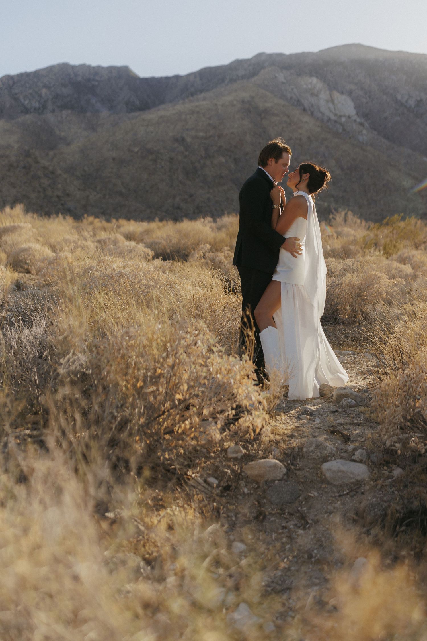 Bride and groom embrace on a desert trail surrounded by dry brush with scenic mountain backdrop at sunset.