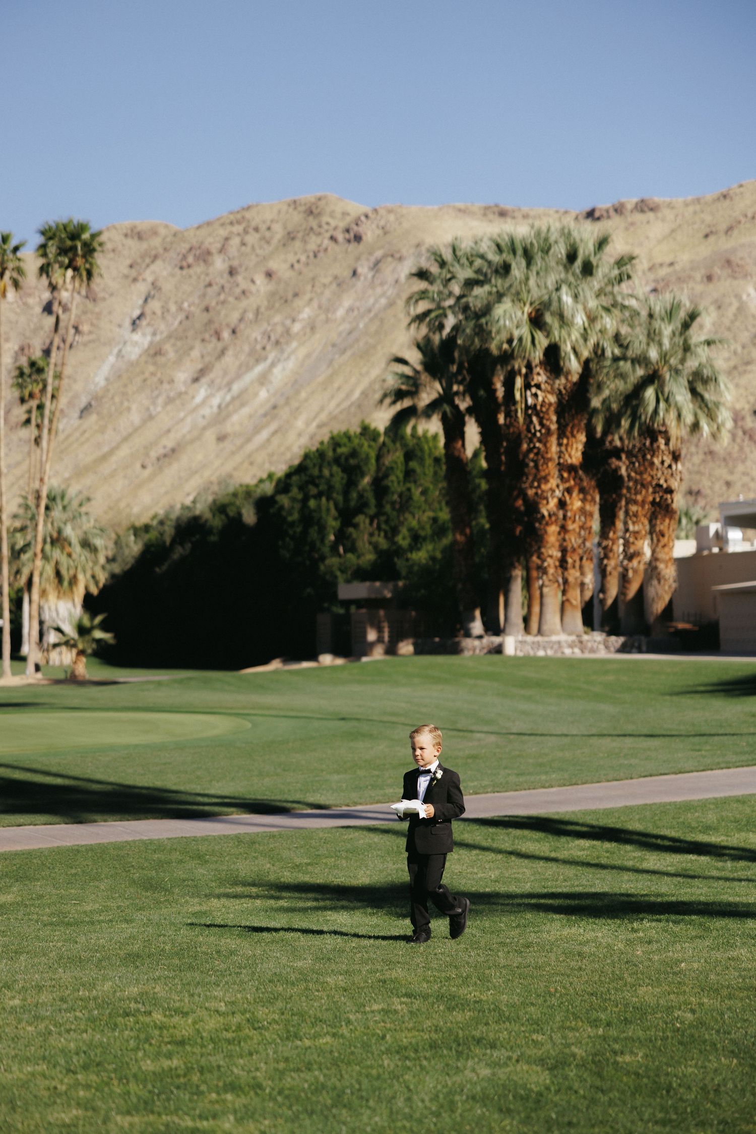Young boy in black tuxedo walking across green golf course with palm trees and mountains.