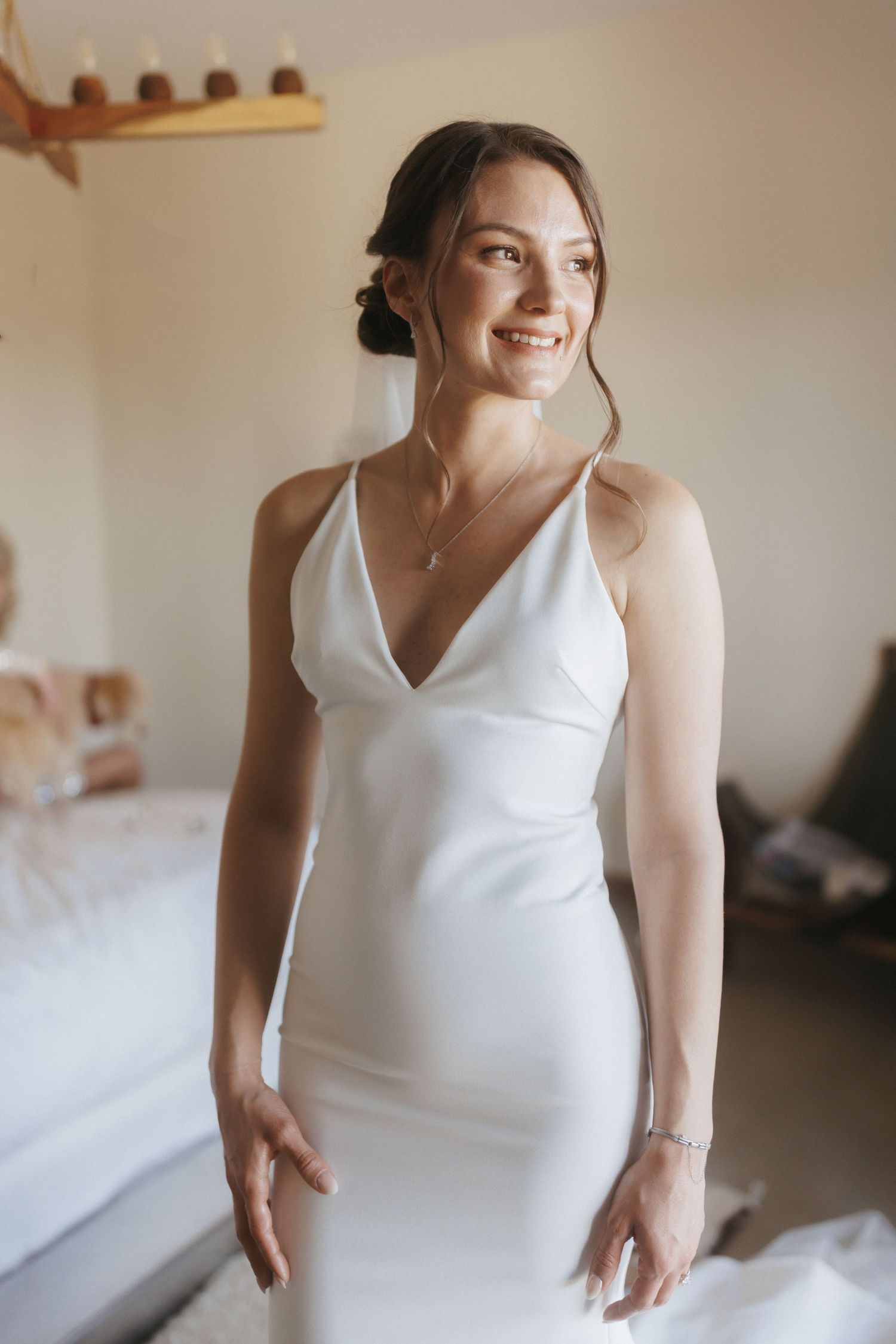 Smiling bride in sleek white satin halter wedding gown with V-neckline and delicate necklace in a bright room.