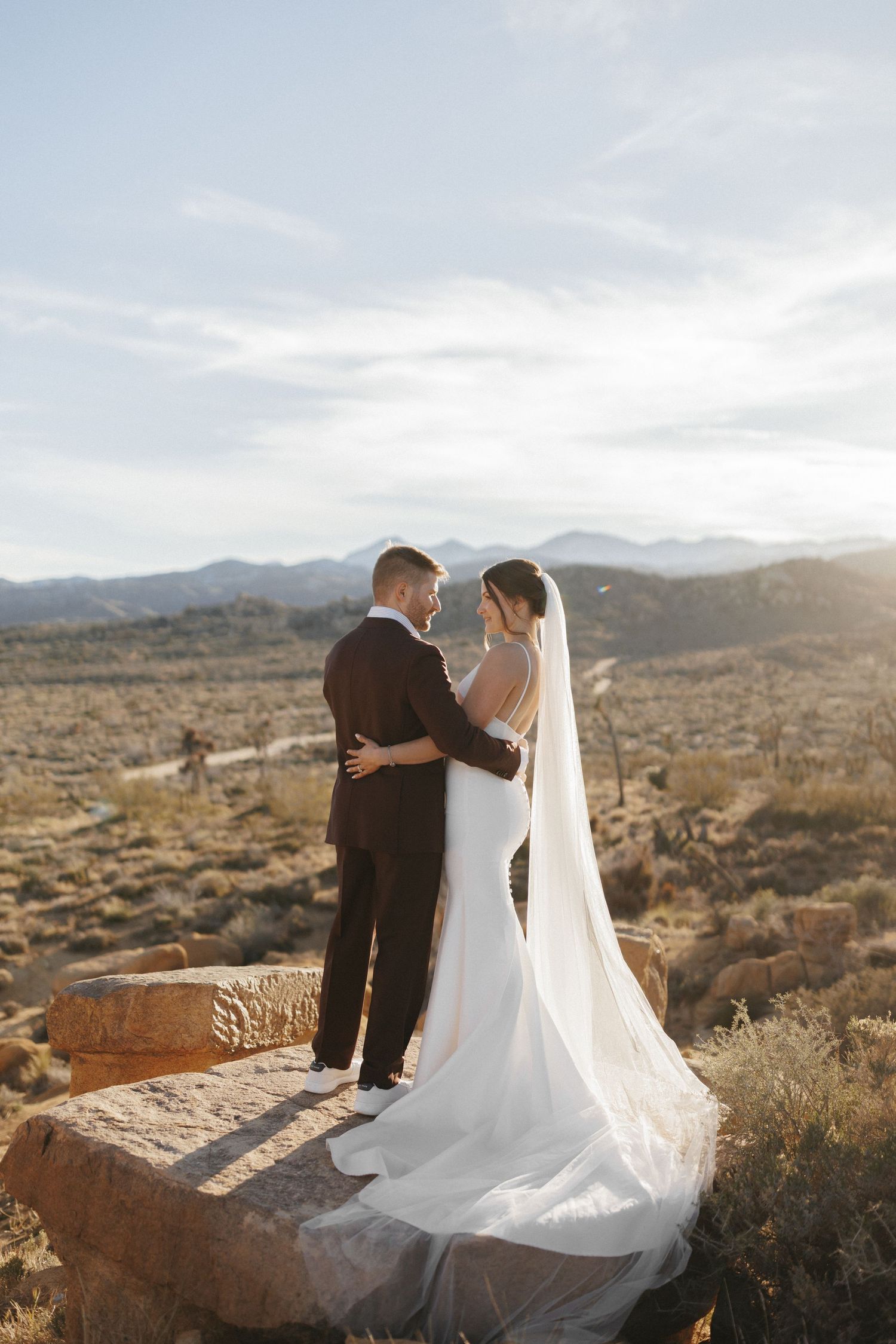 Bride and groom embrace on desert rock with mountain views and flowing veil at golden hour.