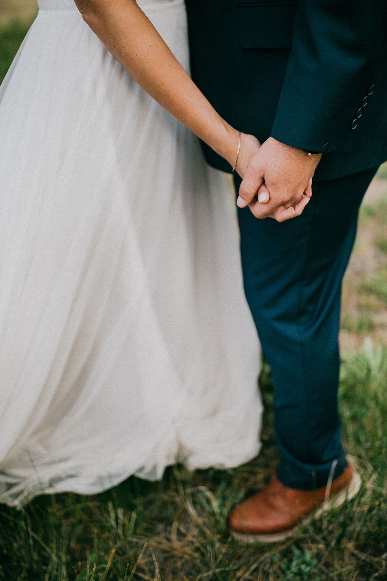 Close-up shot of wedding attire showing white dress fabric and navy suit pants with brown leather shoes on grass.