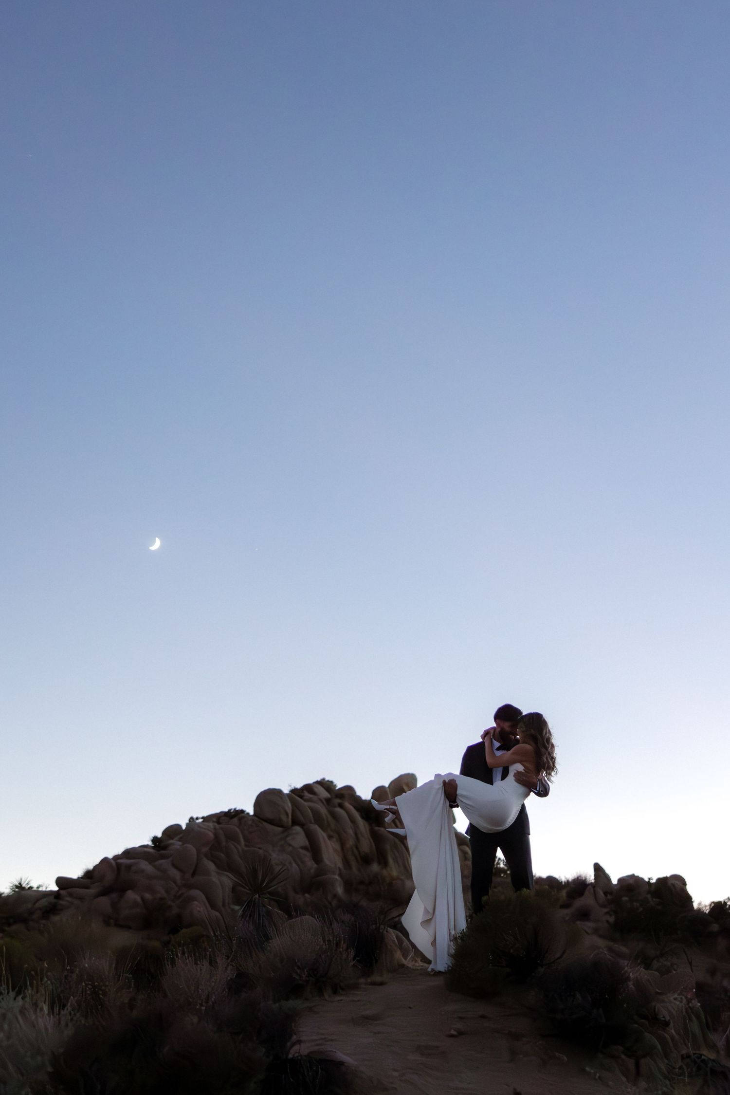 Four silhouetted figures climb rocky terrain against a dusky blue sky.