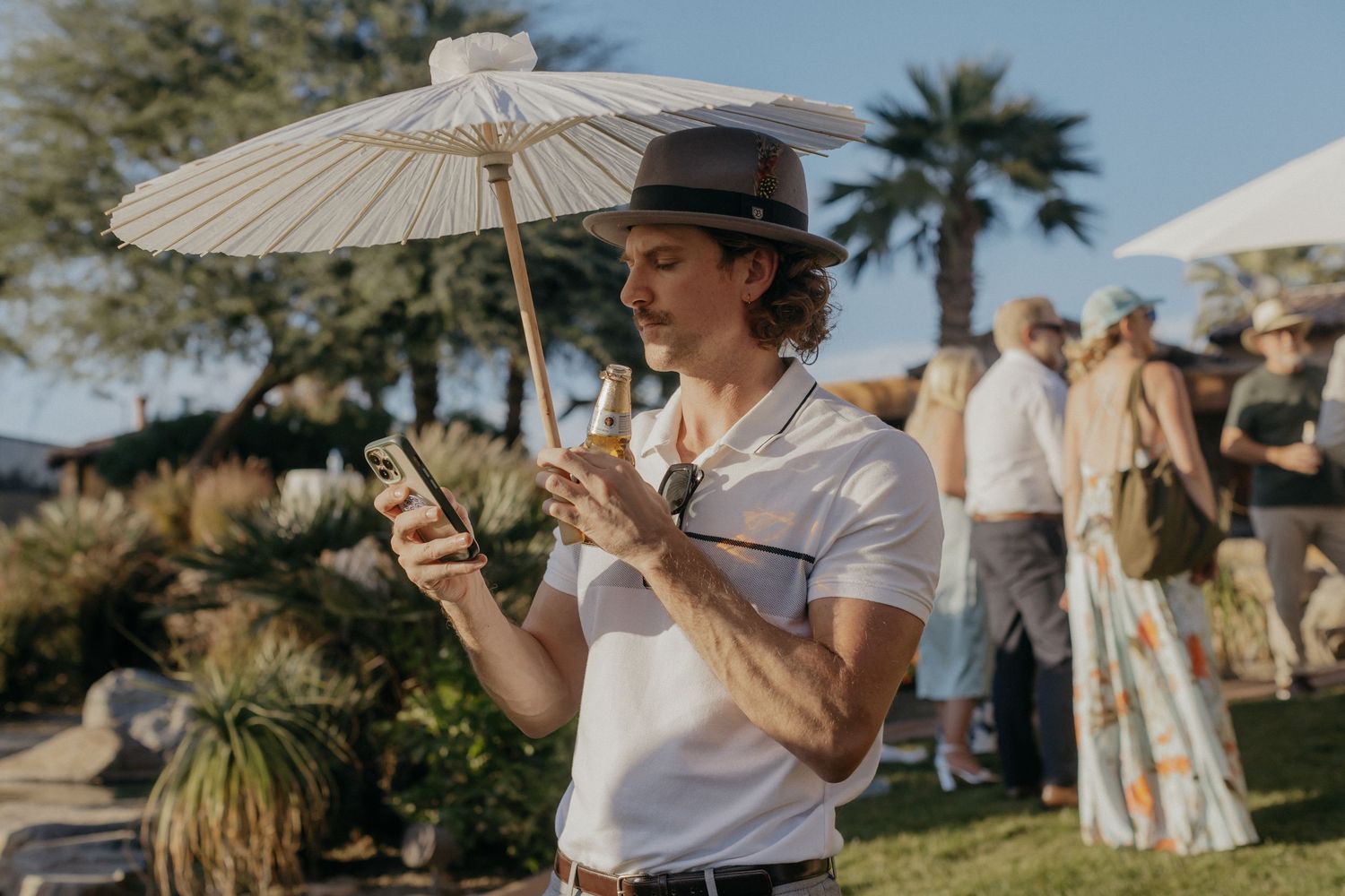 Wedding guests enjoy an outdoor celebration with white parasols and palm trees in the desert backdrop.