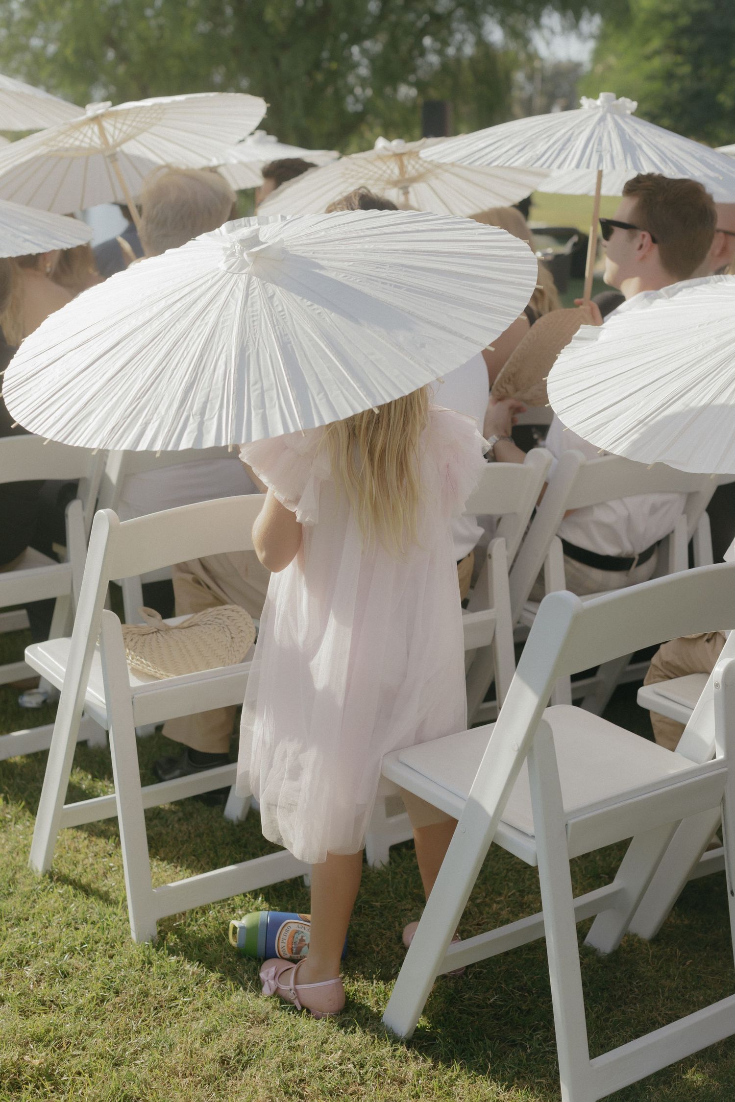Row of white paper parasols providing shade for guests seated on folding chairs at an outdoor event on grass.