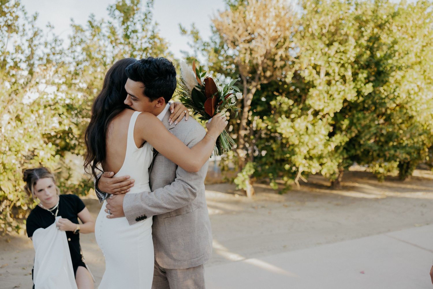 A wedding party shares sweet moments in an outdoor setting surrounded by trees and natural sunlight.
