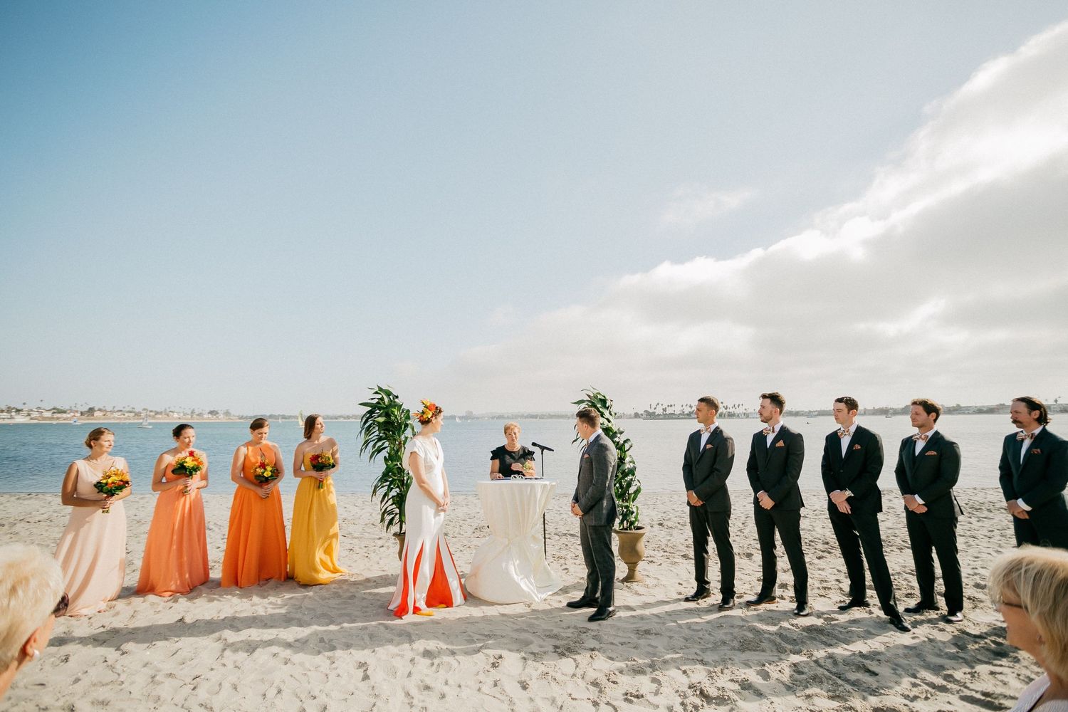 Two groups of wedding party members standing on opposite sides of a beach at sunset, wearing formal attire.