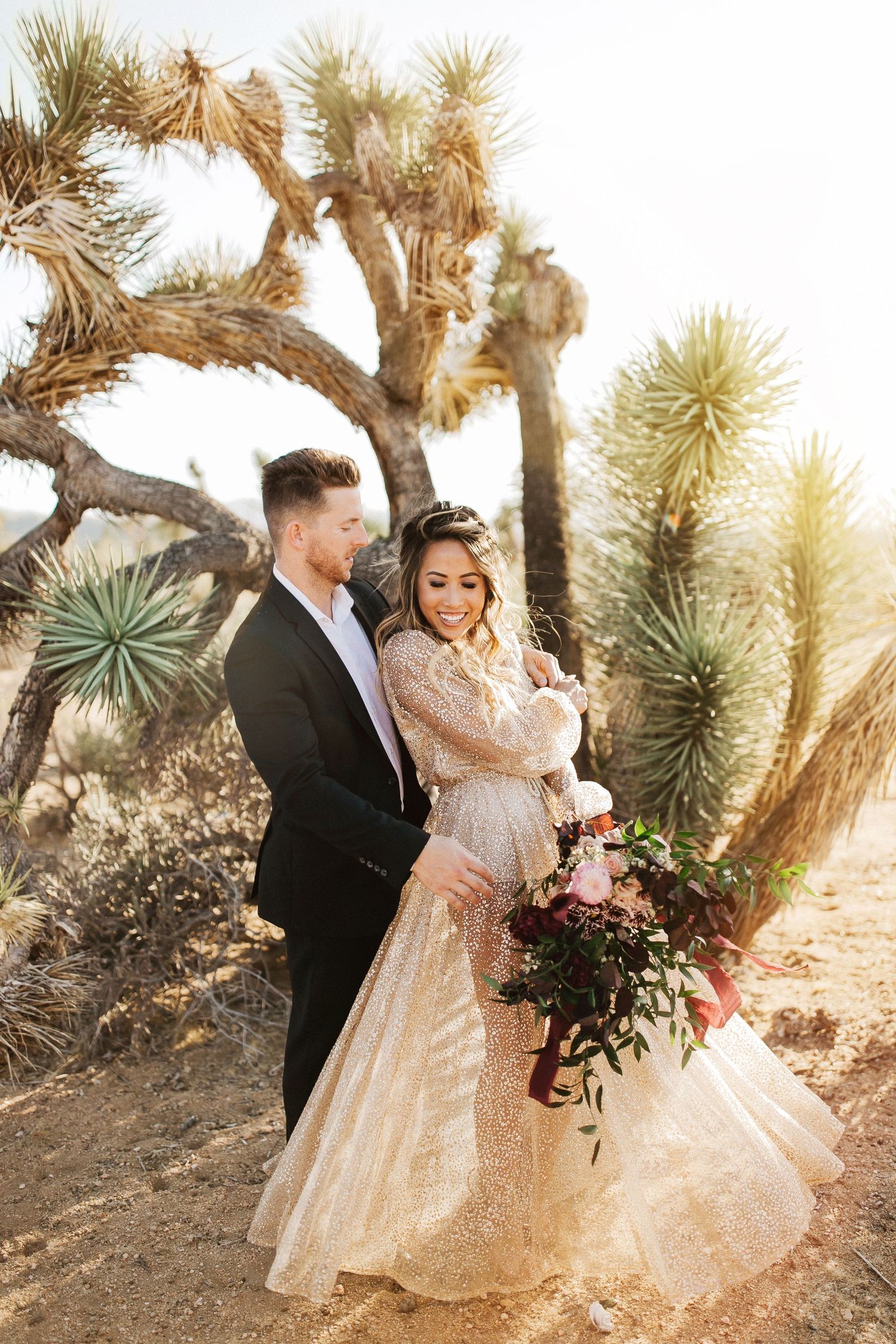 A couple poses in a desert garden with Joshua trees and golden sunlight during their wedding photoshoot.
