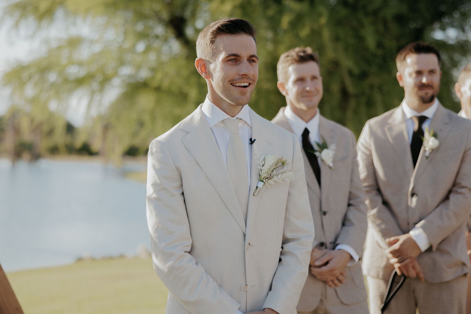 Groomsmen in light beige suits pose together outdoors during a summer wedding celebration.