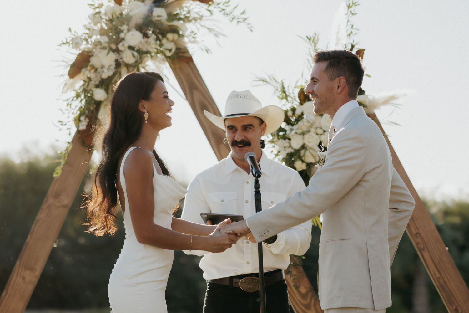 Scenic wedding ceremony takes place under wooden arch during golden hour at sunset.