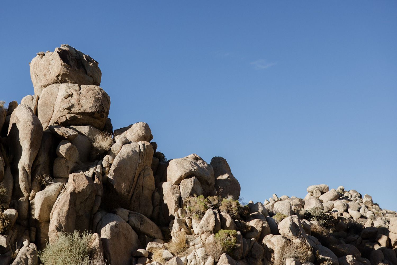 Large granite boulders scattered across a desert landscape against a bright blue sky in Joshua Tree National Park.