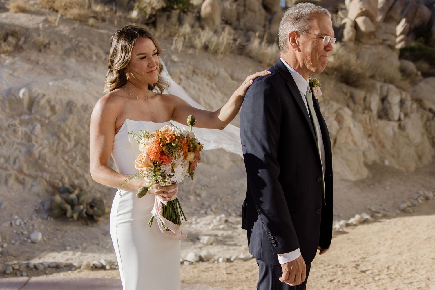 Two people in wedding attire embrace during an outdoor ceremony on a beach at sunset.