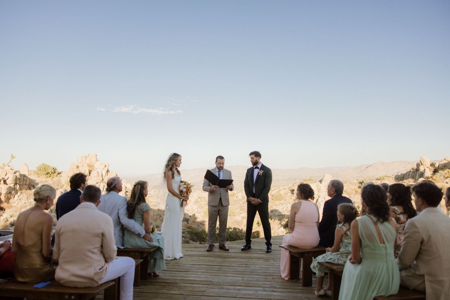 A group of people gathered outdoors at sunset for a wedding ceremony along a wooden boardwalk.