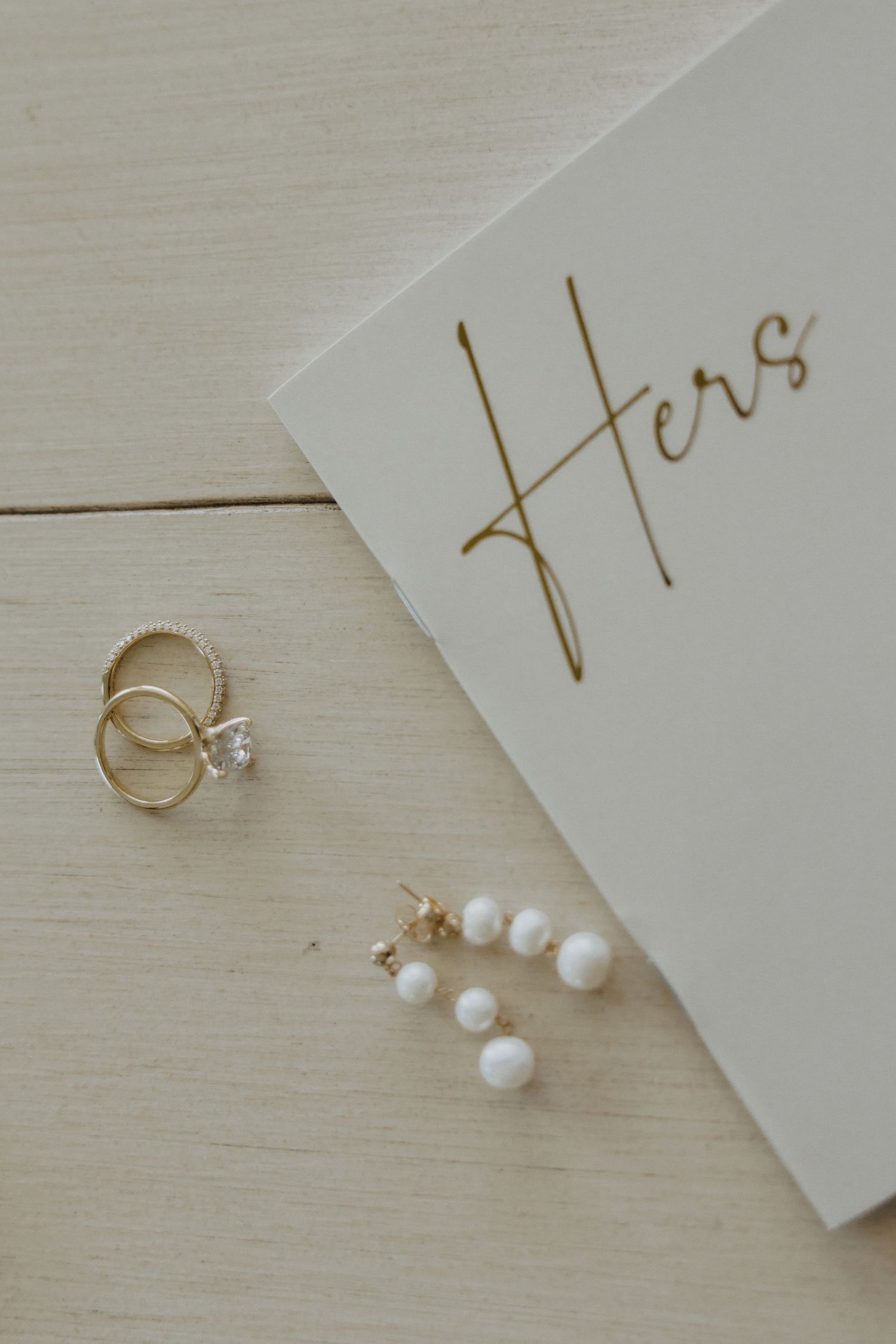 Gold rings and pearl drop earrings beside a white card labeled Hers on a light wood surface.