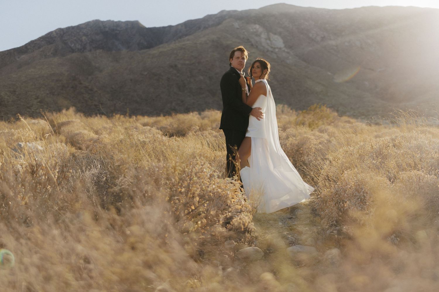 Bride and groom embrace in golden desert landscape with mountain backdrop, bride in flowing white gown with veil.