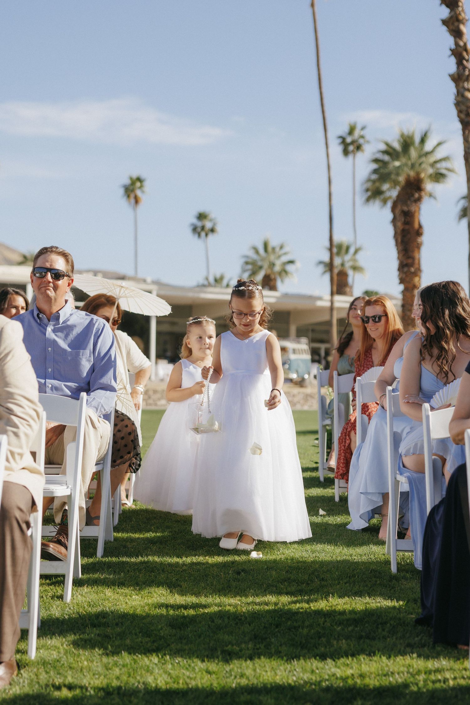 Two flower girls in white dresses walk down the aisle at an outdoor Palm Springs wedding ceremony.
