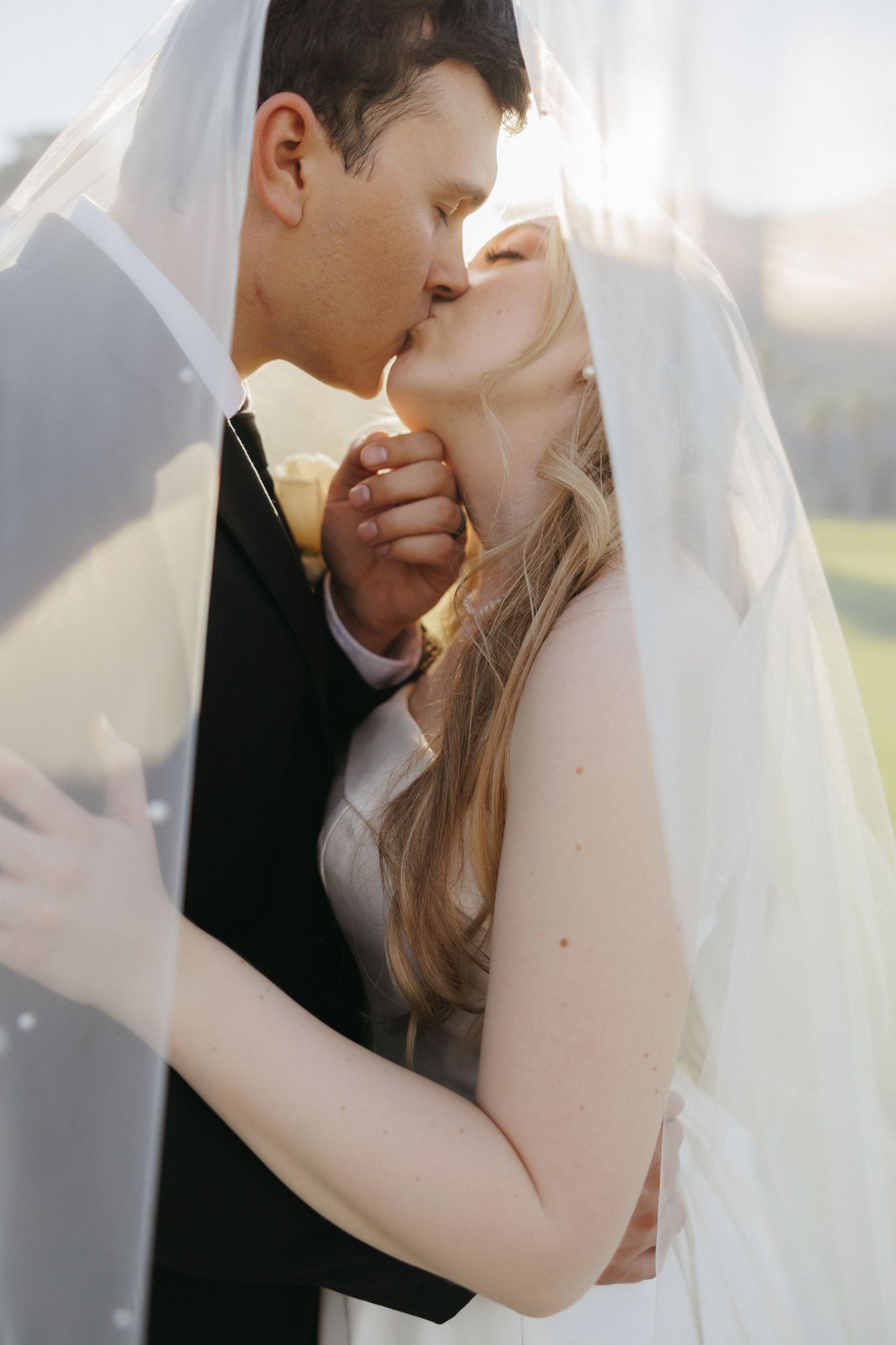 Bride and groom sharing romantic kiss under sheer wedding veil in golden sunlight.