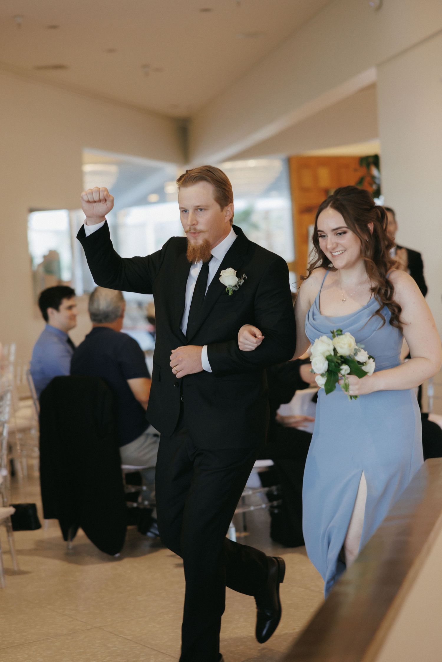 Groomsman in black suit pumps fist while entering reception with bridesmaid in blue dress.