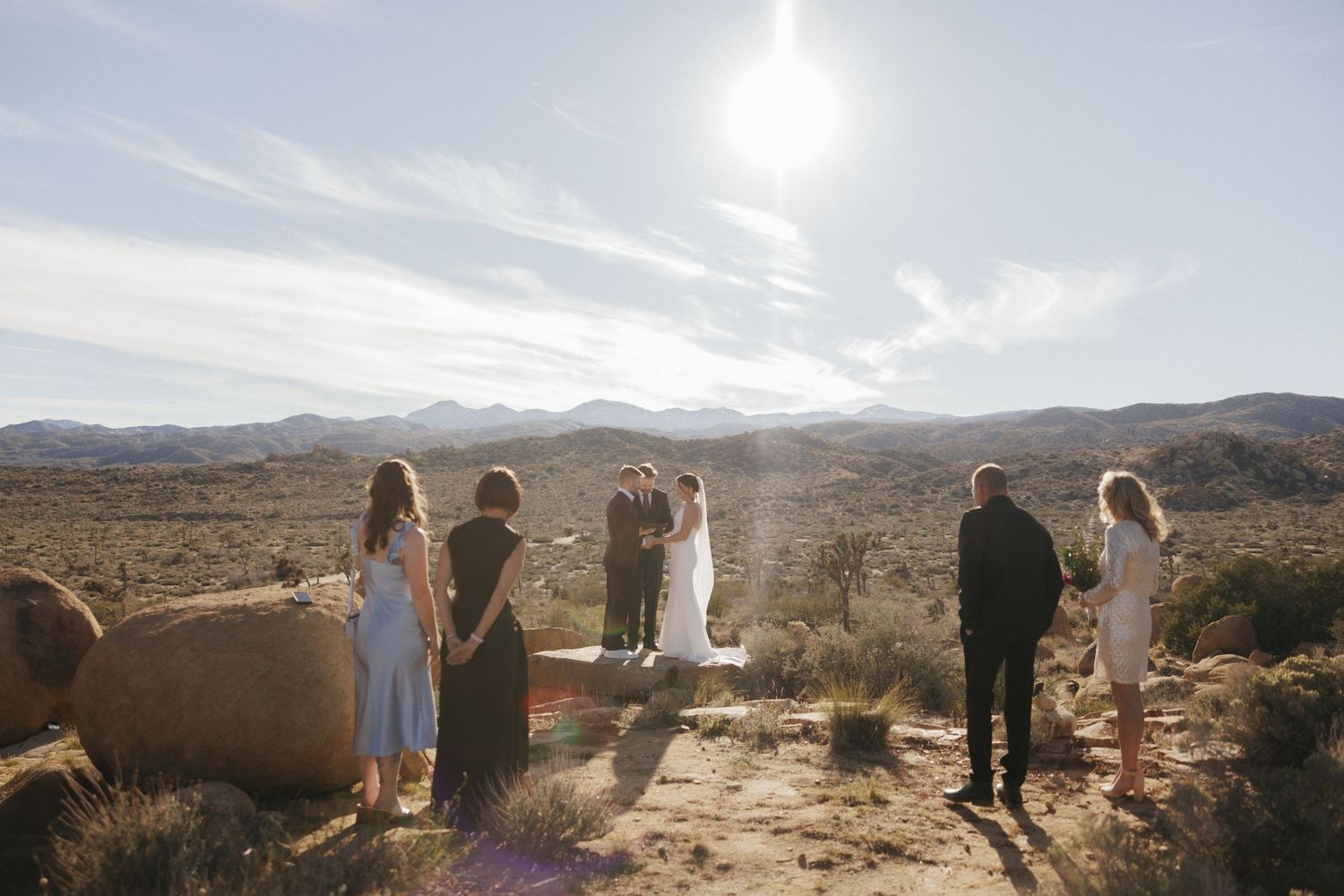 Wedding party gathered for outdoor desert ceremony under bright sun with mountain landscape backdrop.
