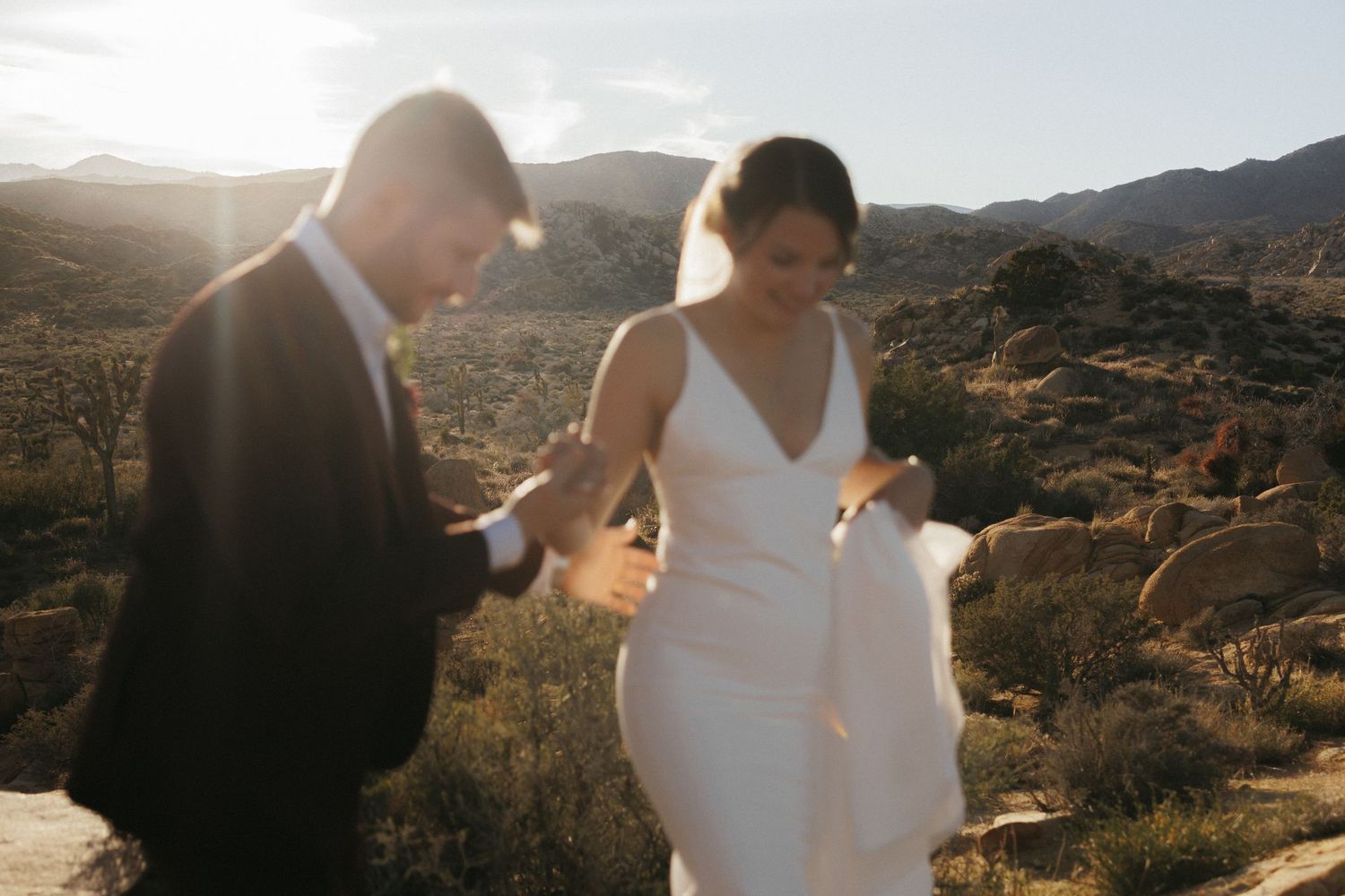 Bride and groom walking together in sunlit desert with mountains in background.