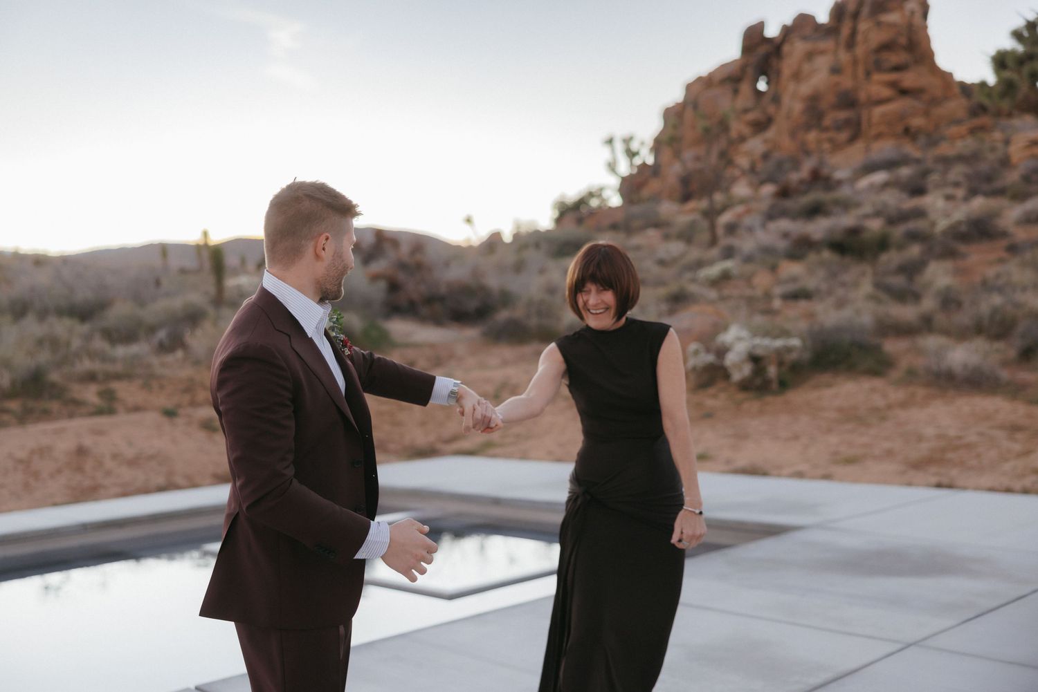 Groom in burgundy suit takes hand of woman in black dress near a reflective pool with desert rock formations at dusk.