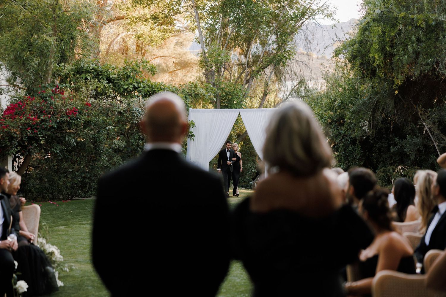 Outdoor wedding ceremony in a lush garden with guests seated, couple at a white arch altar surrounded by greenery.