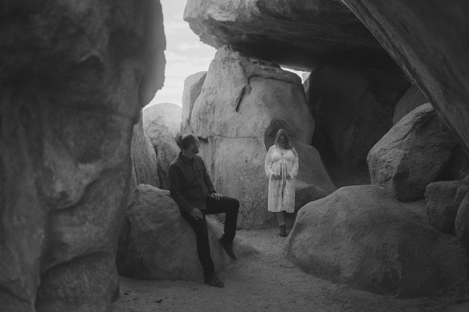 Black and white photo of two people among large rock formations in a desert landscape, one seated wearing a cowboy hat.