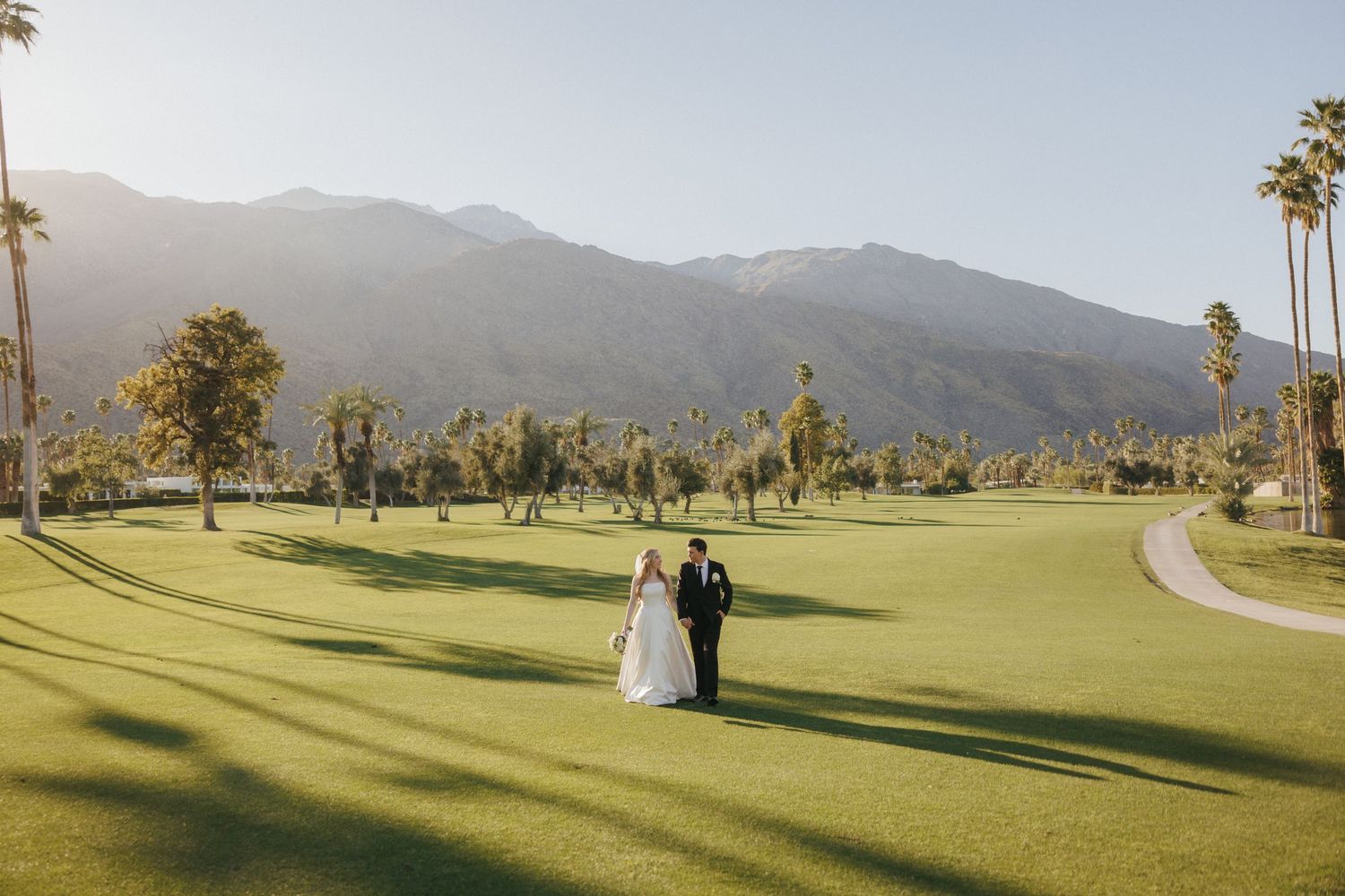 Bride and groom walk hand in hand across green golf course with palm trees and mountains.